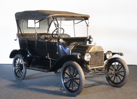 A black 1910s-era car with an open-top carriage and a convertible roof-covering which attaches to the front windscreen and the rear seat. The wheels of the car are very narrow yet have a wide diameter giving the car a high-set seating carriage. 